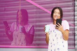 Smiling trendy Chinese woman walking outside in coveralls holding coffee and talking on smart phone