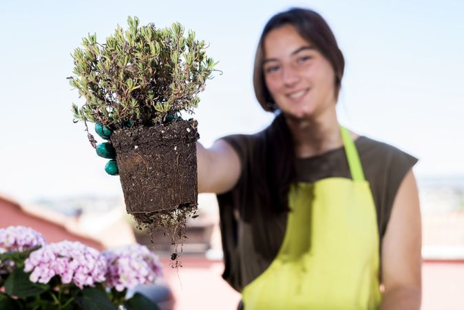 Smiling young woman wearing a gardener apron showing a plant at home terrace