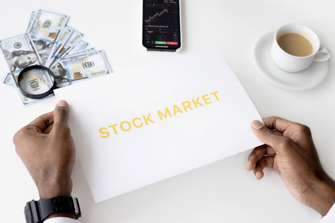 Cropped image of person holding a paper with "Stock Market" written on it near phone and money