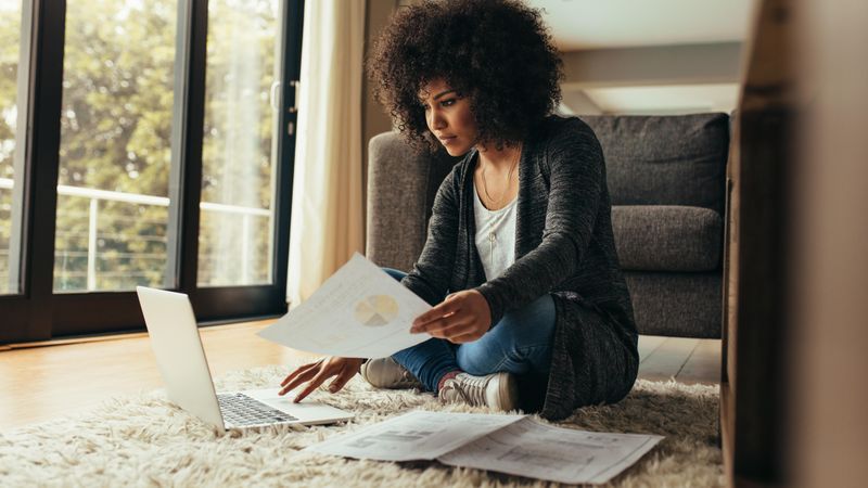 Woman sitting on floor at home with document in hand using laptop