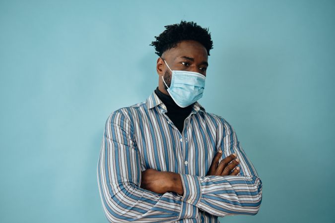 Portrait of a concerned Black man in face mask blue striped shirt with his arms crossed