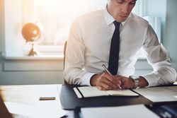 Serious man in shirt and tie sitting at desk and writing in sunny office