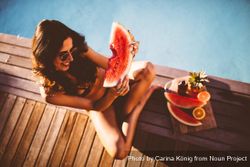 Smiling young woman sitting poolside with a fruit platter eating a slice of watermelon v4Nk8b