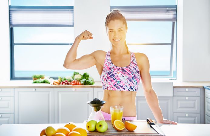 Woman flexing her arm muscle in bright kitchen in front of cutting board with fruit