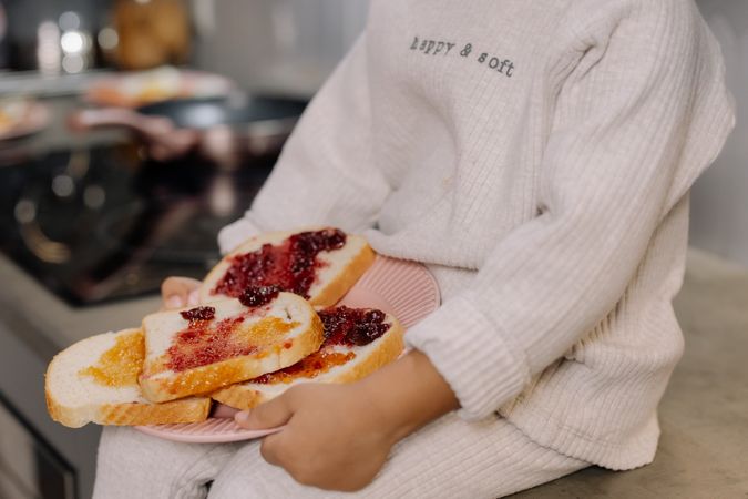Cropped image of a child holding a plate of toasts and jam sitting on kitchen counter