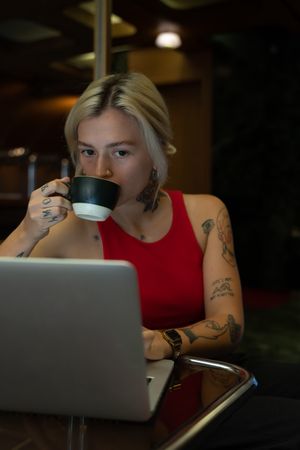Tattooed woman in red top drinking coffee while working on her laptop