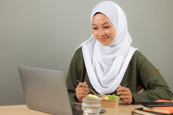 A Beautiful Asian Muslim Woman Wearing a Hijab Enjoys Eating Healthy Yummy Salad Bowl While using her Laptop.