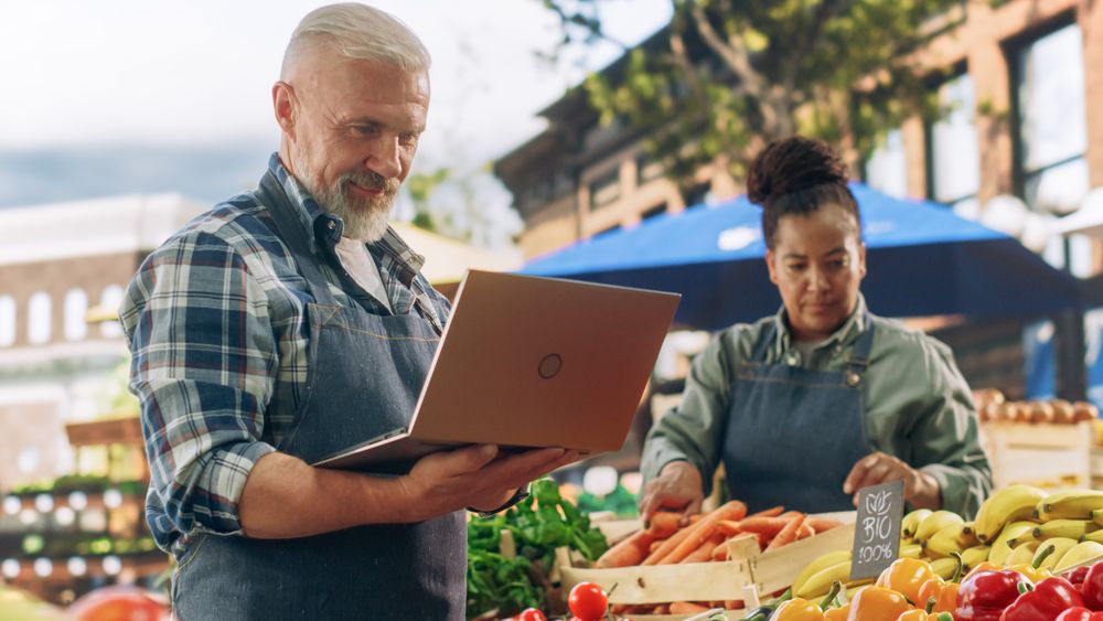 Market Manager Using Laptop Computer while Standing Next to Street Vendor Stalls with Fresh Organic Agricultural Products.