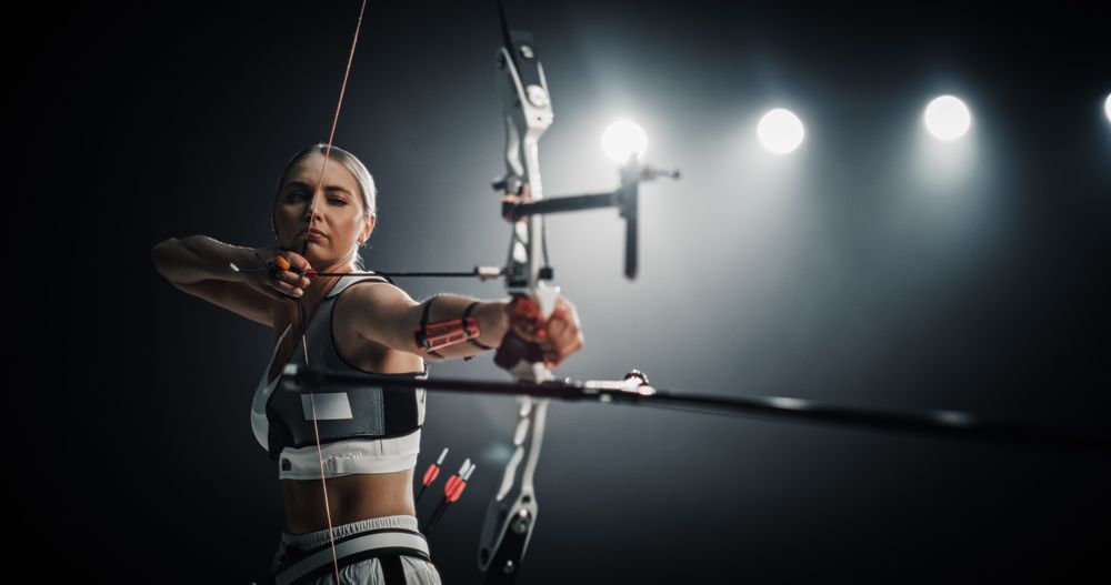 Portrait of Professional Female Archer Aiming an Arrow at a Target with a Compound Bow. Strong Athlete is Fully Equipped and Protected with Chest and Arm Guards, Preparing For Tournament