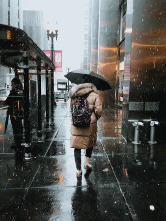 Back view of a person with backpack holding umbrella walking on wet road during rainy day