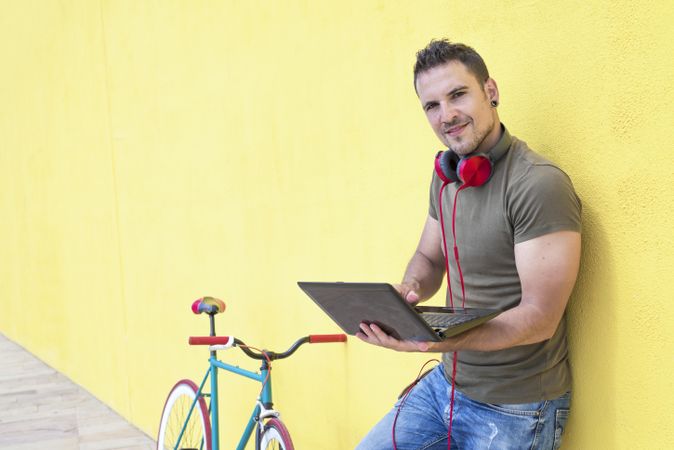 Happy male with red headphones standing outside with laptop and bicycle