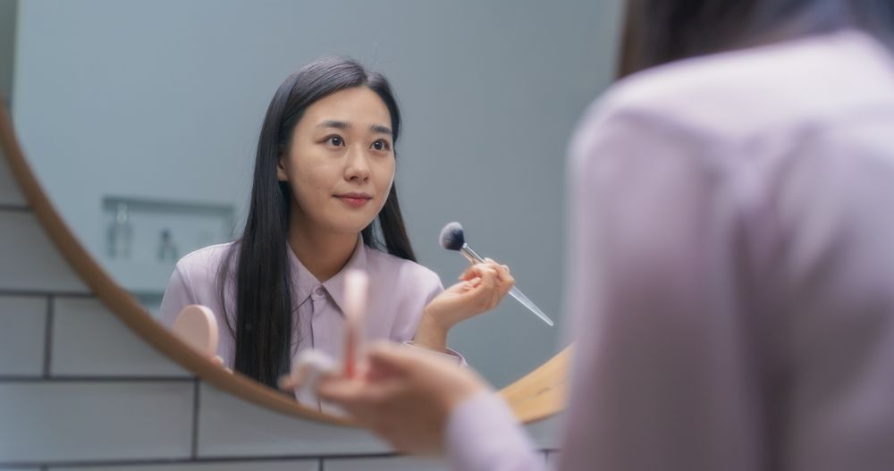 Stylish South Korean Female Using a Soft Brush to Put Tonal Foundation Cream in a Bathroom at Home.