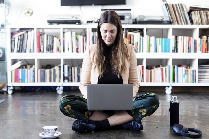 Portrait of a smiling woman sitting cross legged in front of book shelf working on laptop