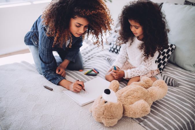 Little girl and her mother sitting on a bed drawing with crayons in a sketch book