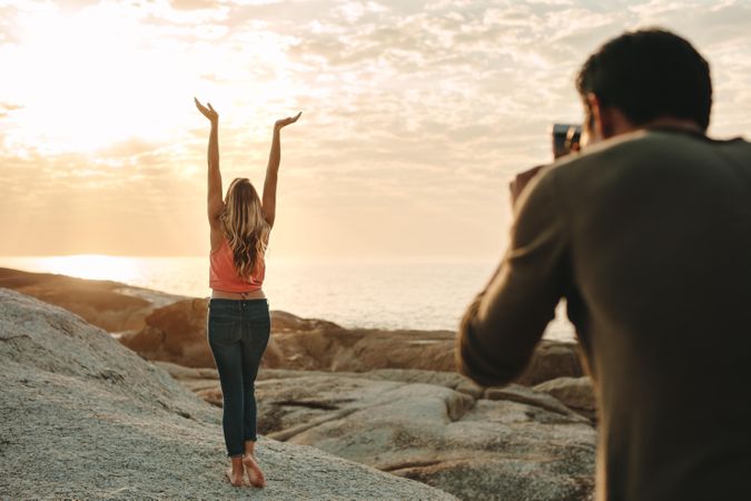 Man taking photo of a woman standing on a rock at the beach using a mobile phone