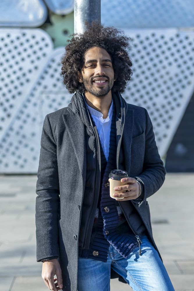 Happy Black man in jacket with warm beverage standing outside leaning on pole