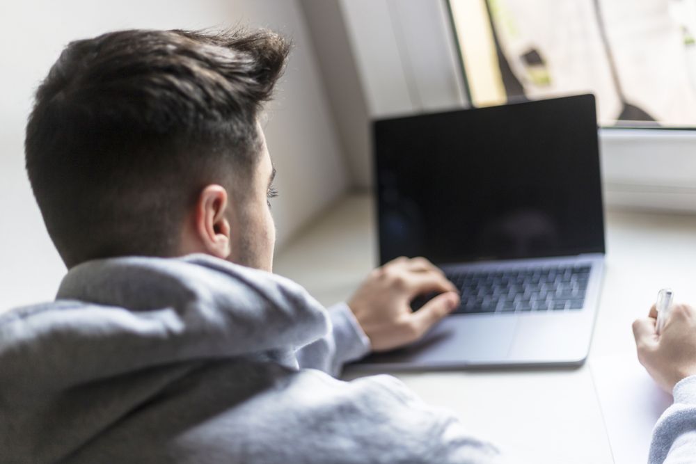 Rear view of a young man working on laptop by window at home