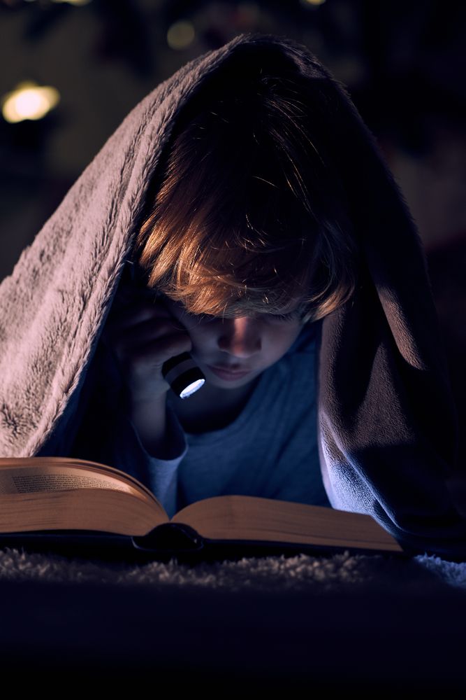 Boy reading reading book in darkness under blanket