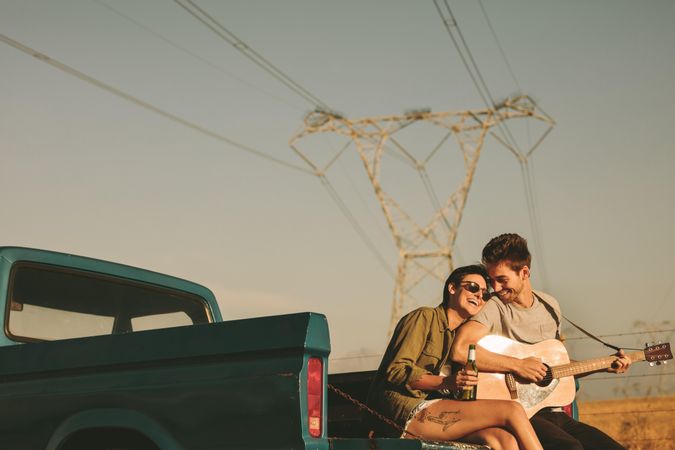 Young woman and man sitting on edge of pickup truck playing guitar while parked on country road
