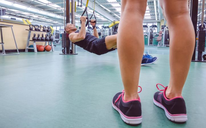 Legs of trainer looking on at person using calisthenics in gym