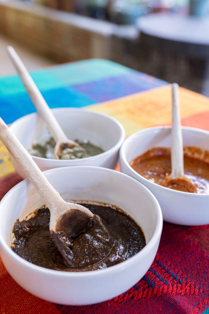 Three bowls with red, green and yellow mole sauce with a wooden spoon on top of a colorful traditional tablecloth