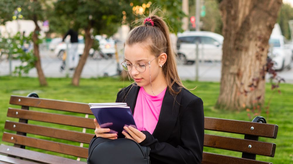 Girl reading book while sitting on park bench.