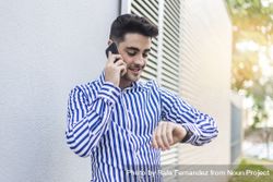 Young man standing outdoors and checking his wrist watch while speaking on phone 5XrYEV