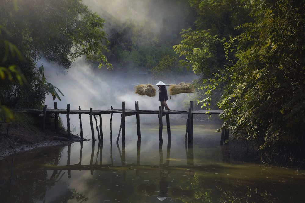 Person wearing conical hat walking on bridge over lake