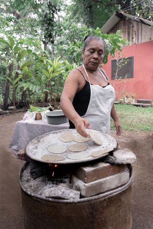 A Hispanic woman is cooking traditional corn tortillas over an outdoor stove