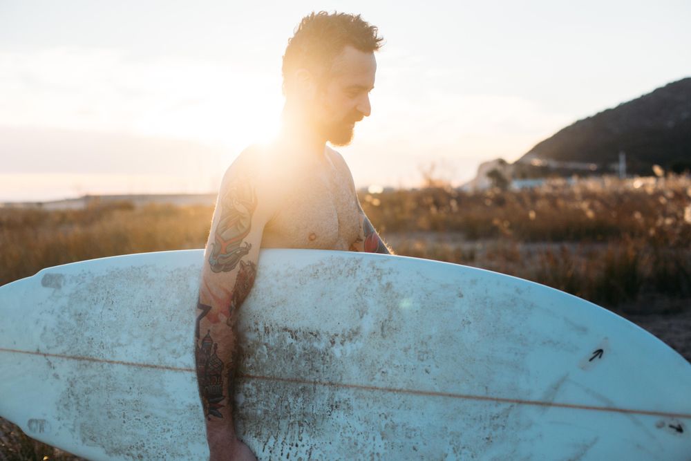 Tattooed man looking down with surfboard under his arm with sun behind him