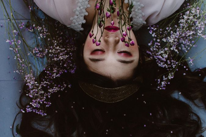Upside down photo of woman with pink makeup lying on the ground next to pink flowers