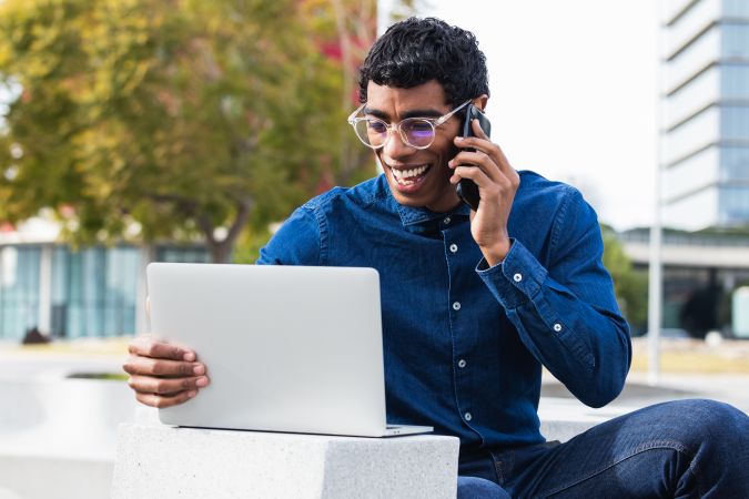 Happy hispanic businessman freelancer working on laptop and talking on mobile phone in city