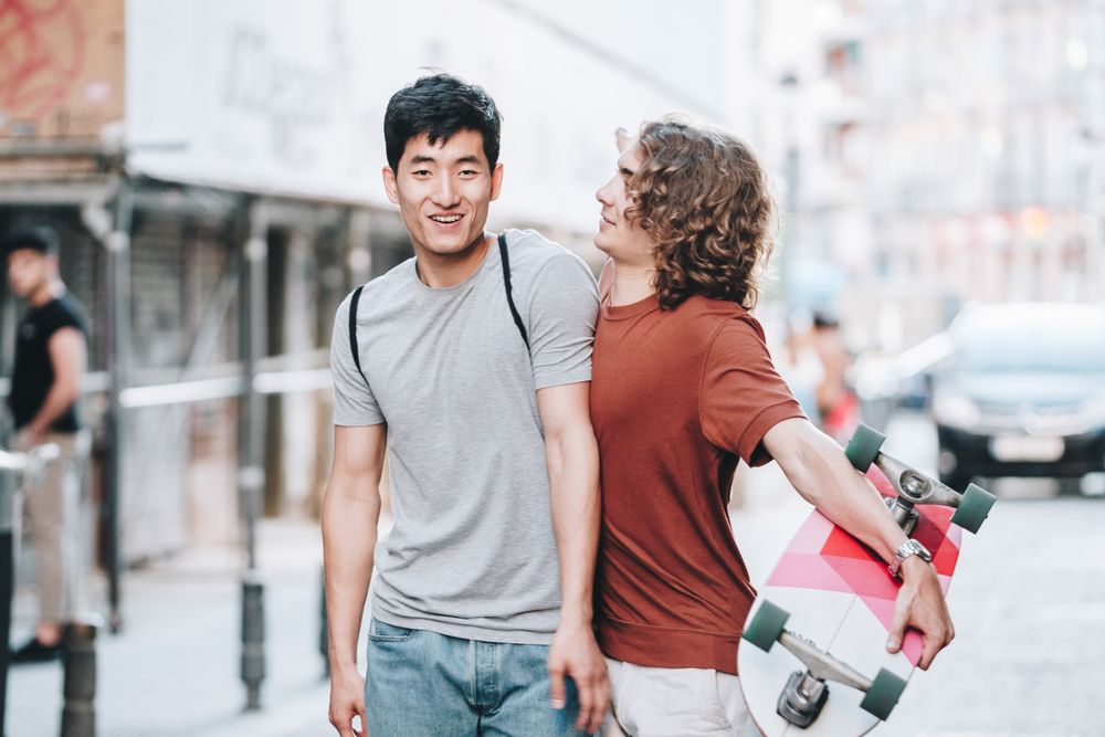 Cool multiracial male friends chatting while walking on street with longboard