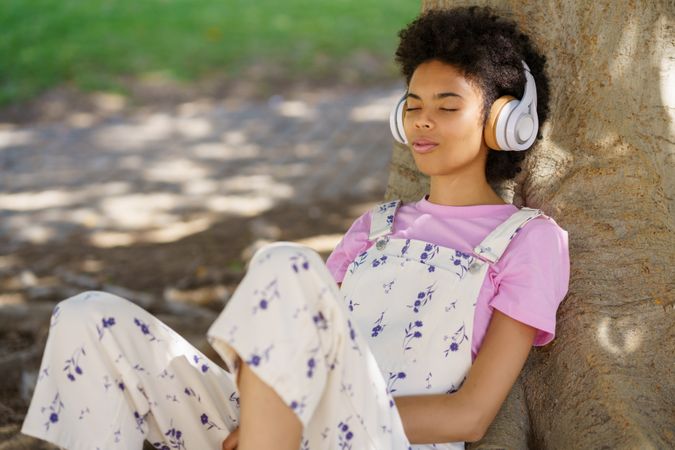 Content woman in floral coveralls relaxing on roots of large tree in park listening to music