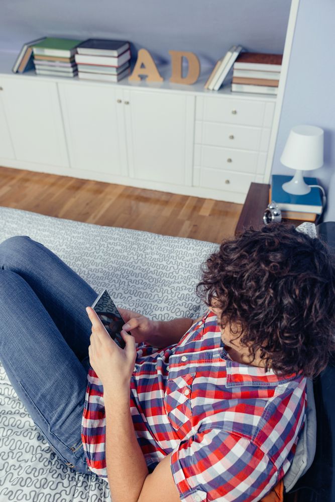 Young man with curly hair looking smartphone in a bed