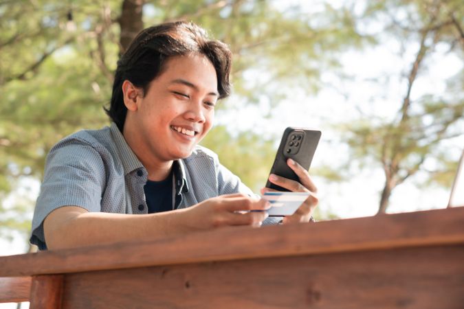 Happy Asian Young businessman hold blue creditcard and phone purchasing online shopping in outdoor cafe.