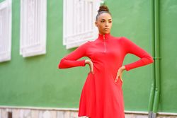 Serious female in red zipped up dress standing outside green building