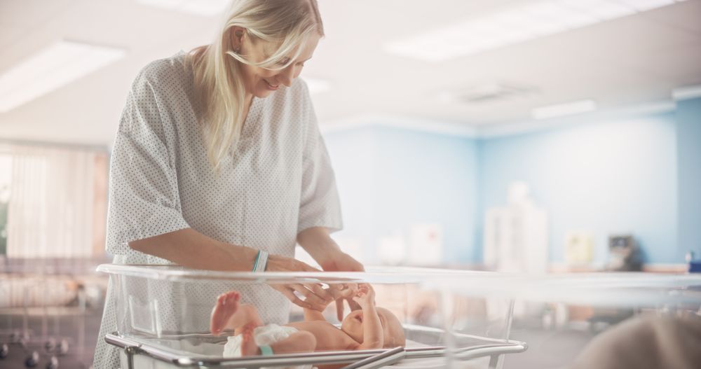 Mother Bonding with her Newborn Child Near Hospital Bassinet Bed in a Modern Nursery Clinic.