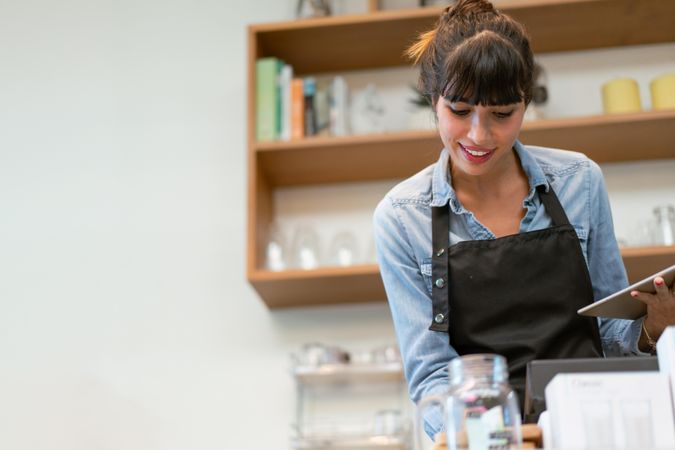 Smiling female at coffee shop counter wearing apron with tablet
