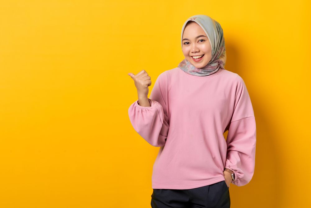 Smiling Muslim woman in scarf and pink sweater pointing to side with thumb