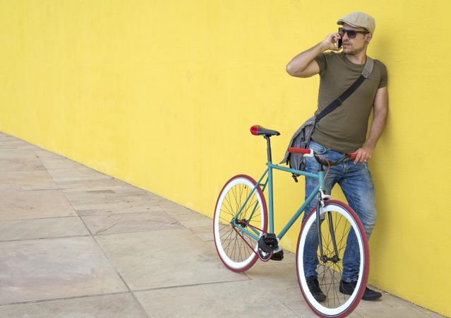 Male in hat and sunglasses leaning on yellow wall with bike and taking call on phone
