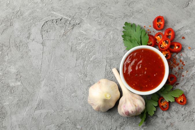 Bowl with chilli sauce, pepper and garlic on gray background