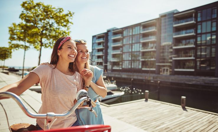 Two happy female friends laughing while walking along a riverwalk with bike on sunny day, copy space