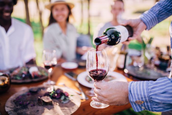 Man pours a glass of red wine at an outdoor table setting among friends