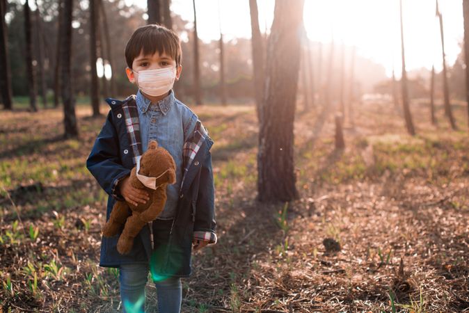 Boy with facemask holding teddy bear near trees