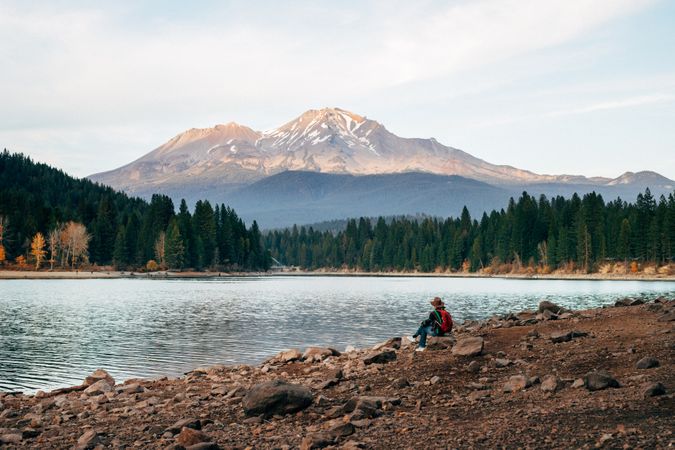 Person with fedora hat and backpack sitting on river shore in woods near Mt. Shasta in Siskiyou County, CA, US