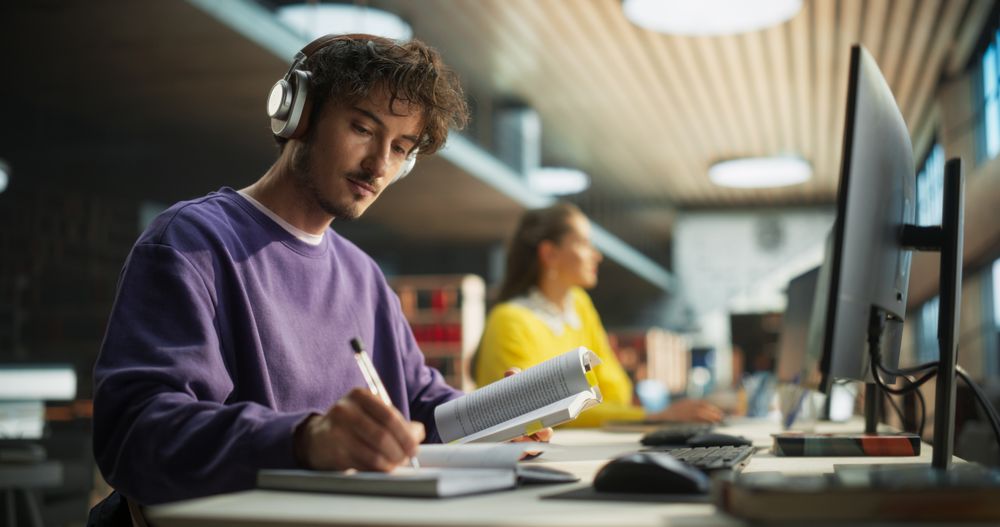 Handsome University Student Studying in a Traditional Public Library with Other Scholars. Young Male Wearing Headphones, Sitting in a Hall, Reading Academic Textbook and Preparing for Exams