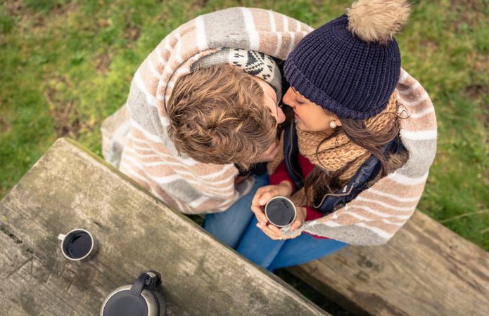 Top view of couple kissing while holding warm coffee outside on park bench