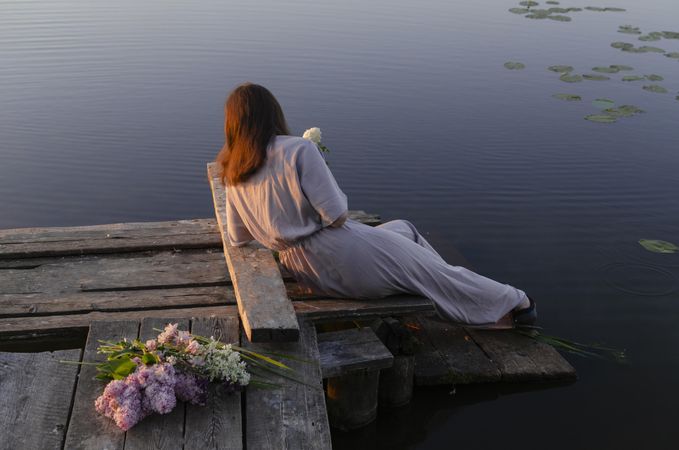 Back view of woman in stylish outfit sitting on wooden dock beside pink flowers
