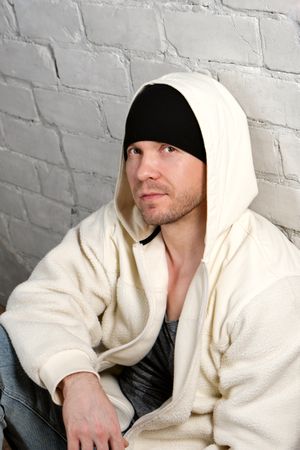 Man in fleece hoodie and beanie sitting against wall.